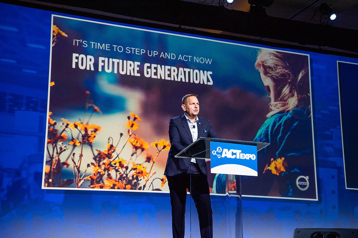 man addressing audience at podium with video screen in background
